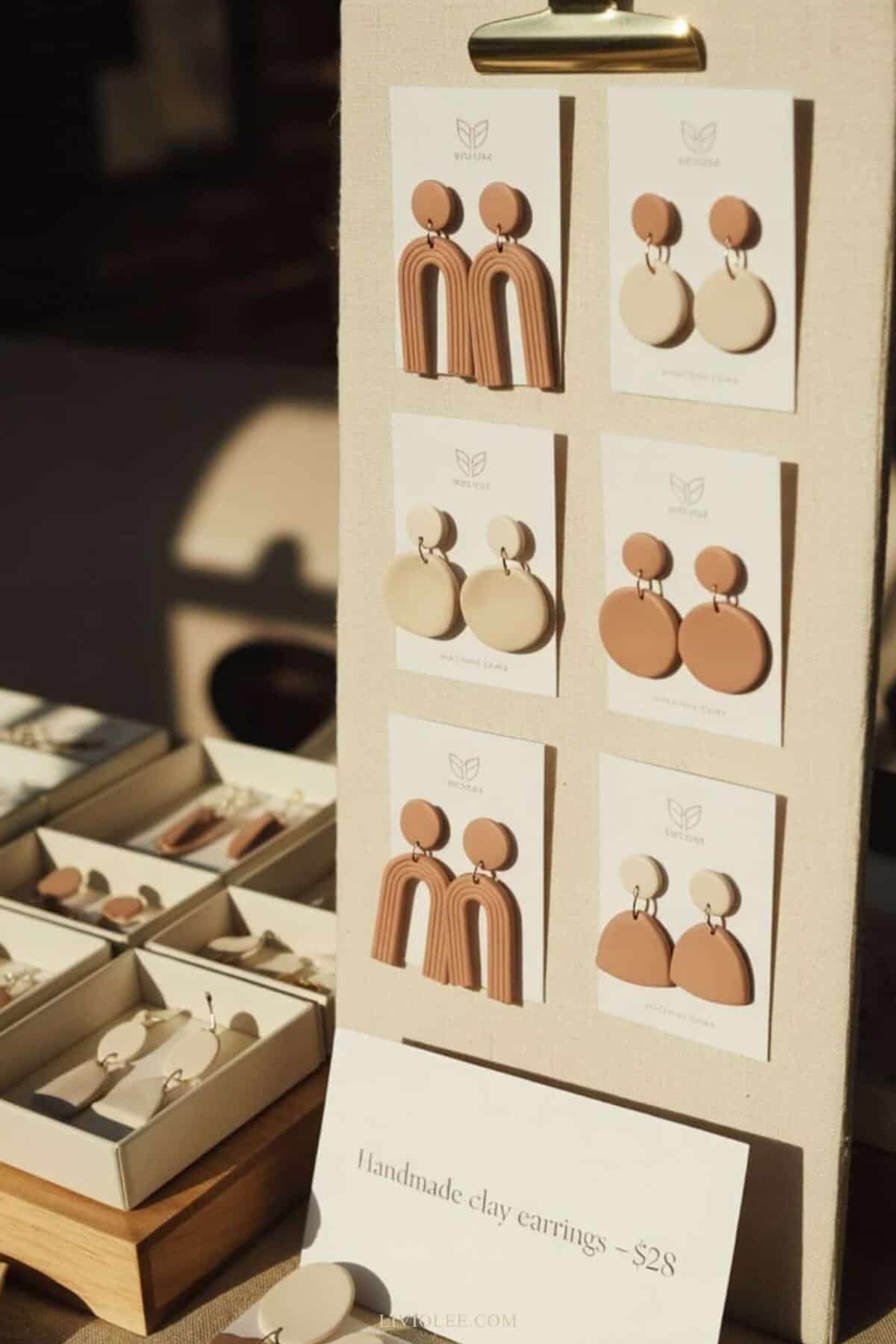 clay earrings at a boutique-style craft market. Neutral-toned earrings (arches, circles, half-moons) mounted on elegant white cardstock with a simple brand logo
