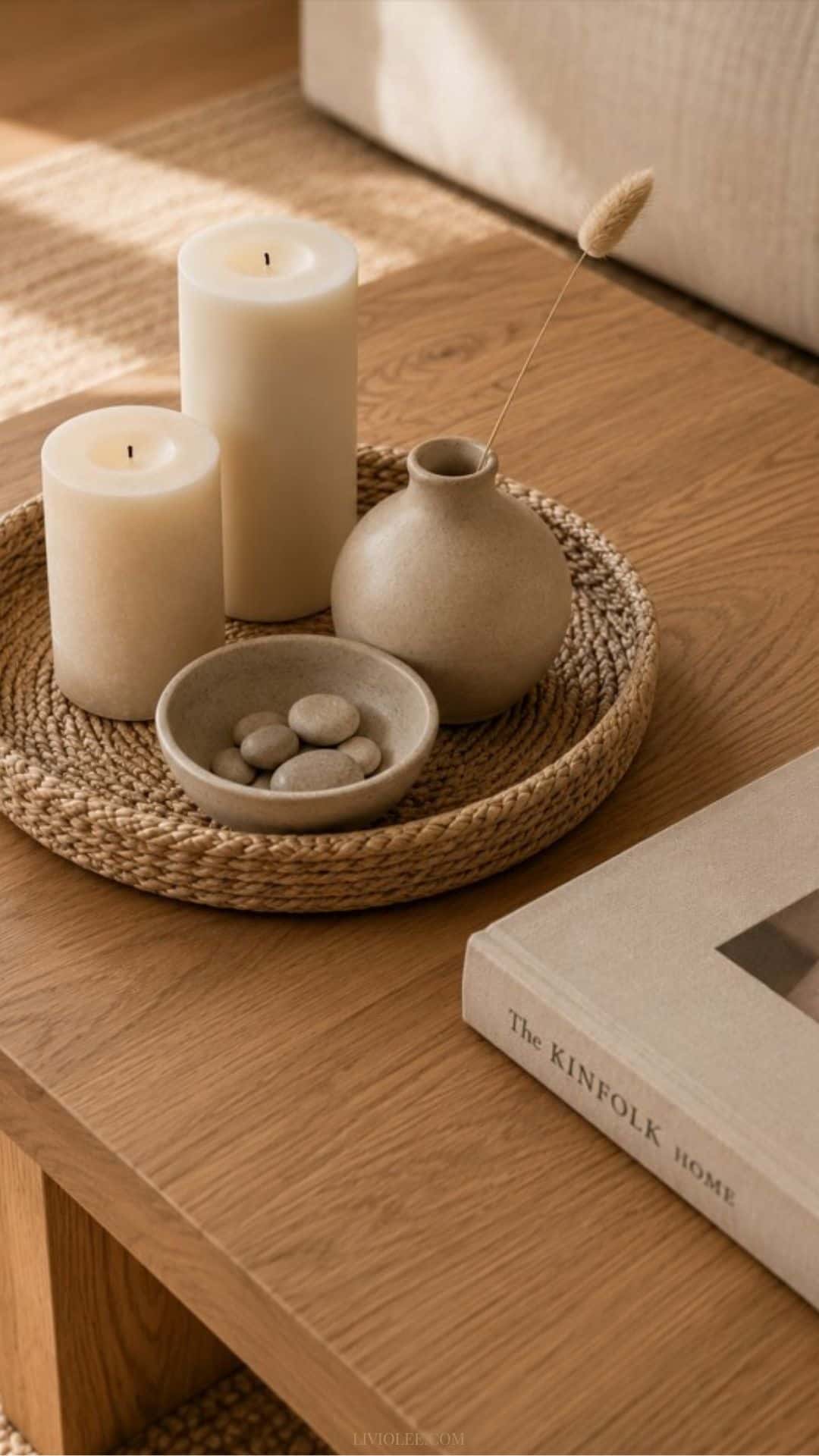warm oak coffee table styled in a minimalist aesthetic with a woven tray holding two cream pillar candles, a matte ceramic bowl filled with smooth stones, and a small ceramic vase with a single dried stem