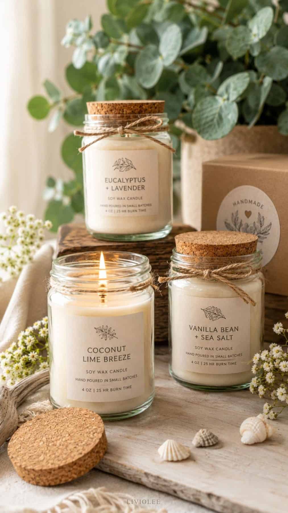 Softly styled recycled glass jar candles made with creamy soy wax, twine ties, and cork lids arranged on a rustic wooden surface with eucalyptus branches, linen fabric, seashells, and delicate white flowers
