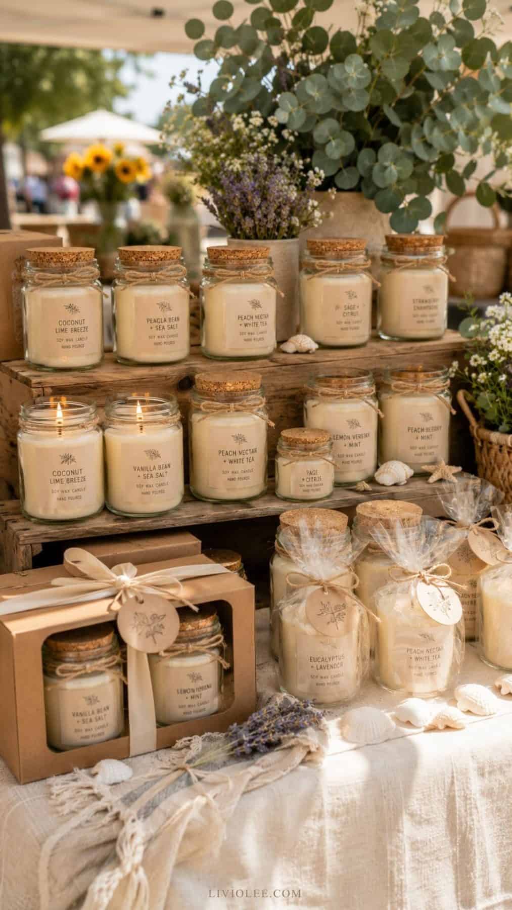 market display featuring handmade recycled glass jar candles in assorted sizes and scents, arranged on wooden risers with eucalyptus, lavender, seashells, and linen table coverings. Some candles are gift wrapped in kraft boxes and clear cellophane with twine and tags